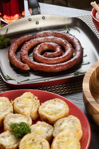 Foto salsiccia e pane all'aglio su un piatto rosso sul tavolo barbecue con antipasti, formaggio, rosmarino, olive e pomodorini.