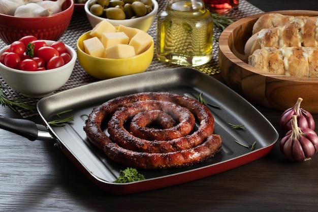 Sausage and garlic bread on a red plate on the barbecue table with appetizers, cheese, rosemary, olives and cherry tomatoes.