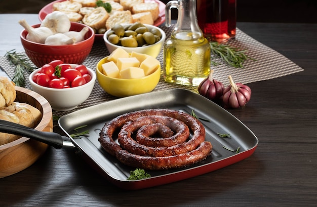 Sausage and garlic bread on a red plate on the barbecue table with appetizers, cheese, rosemary, olives and cherry tomatoes.