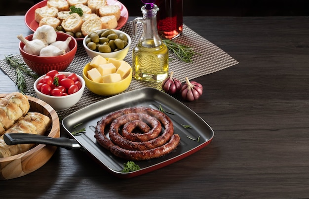 Sausage and garlic bread on a red plate on the barbecue table with appetizers, cheese, rosemary, olives and cherry tomatoes.