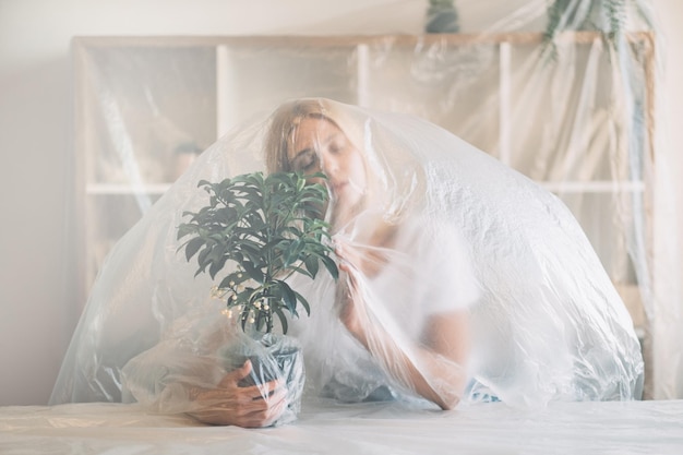 Save nature Defocused silhouette Plastic pollution Ecology problem Caring woman covered with polyethylene film protecting young tree growing in pot at home on light background out of focus