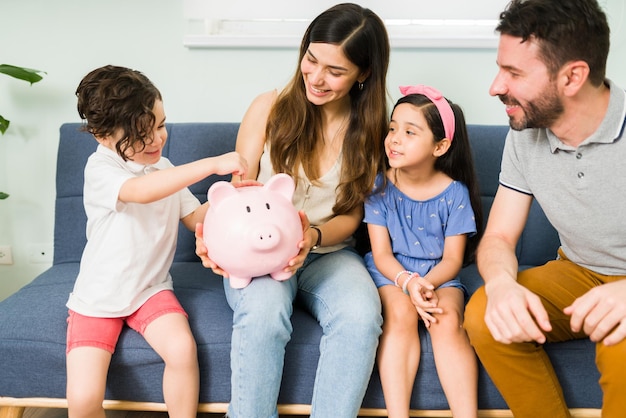 Saving for a family vacation. Beautiful family of four putting money on a small piggy bank while sitting together on the sofa