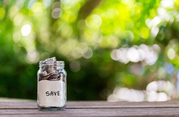 Savings deposit coins in a clear glass bottle, on a wooden floor with bokeh in the backgro