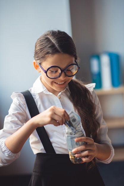 Savings. Girl in white shirt putting money into a jar