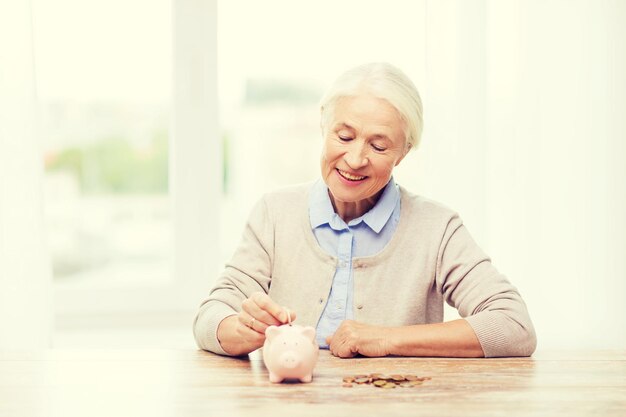 savings, money, annuity insurance, retirement and people concept - smiling senior woman putting coins into piggy bank at home