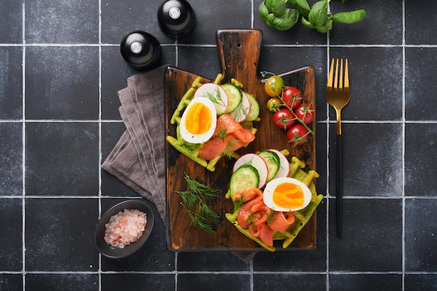짭짤한 와플 아침 식사 녹색 와플 야생 마늘 또는 계란 토마토 연어를 나무 접시에 넣은 시금치 오래된 검은 타일 테이블 배경에서 아침 식사로 가벼운 저녁 점심 또는 아침 식사 상위 뷰