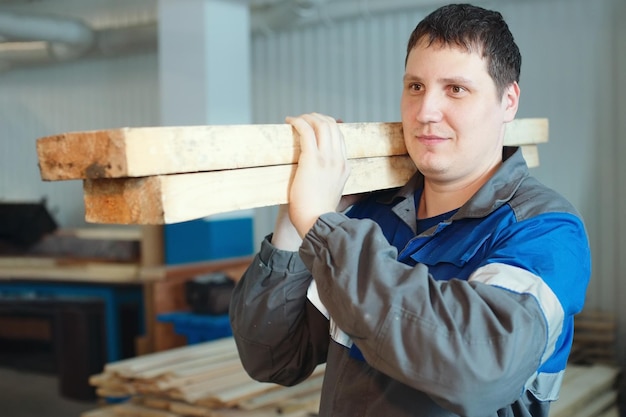 A sawmill worker in overalls carries wooden bars on his shoulder Production and supply of construction materials