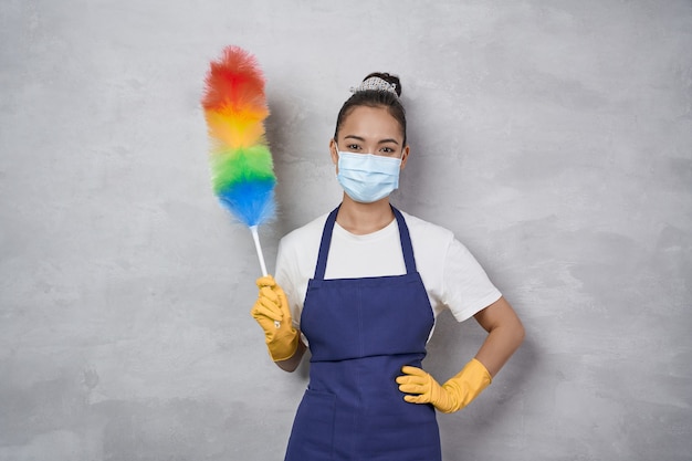 Say No to Dust. Young woman, cleaning lady in uniform wearing medical mask holding colorful microfiber duster and looking at camera while standing against grey wall. Cleaning services during pandemic