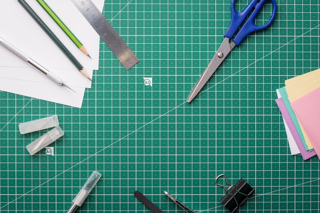 Scalpel, tweezers, ruler, paper clip, box with blades, colored papers, pencils, scissors on cutting mat background