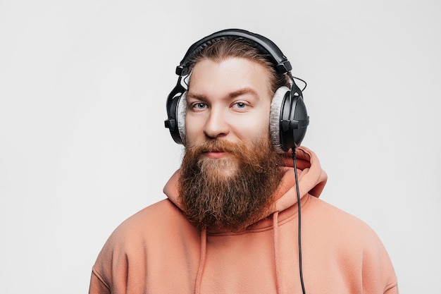 Scandinavian handsome contented man looks smiles and listens to music in professional headphones isolated on gray background Happy guy with ginger hairstyle and beard Modern digital technologies