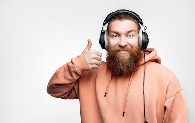 Scandinavian handsome man shows nice thumbs up gesture smiles and listens to music in professional headphones on gray background Happy guy with ginger hairstyle and beard Modern digital technologies