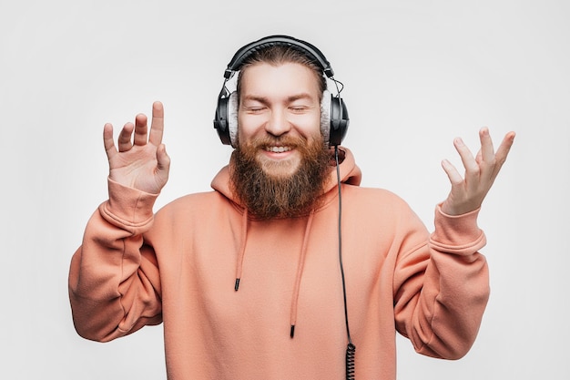 L'uomo sorpreso bello scandinavo ride e ascolta la musica in cuffie professionali isolate su sfondo grigio ragazzo felice con la barba acconciatura allo zenzero tecnologie digitali moderne