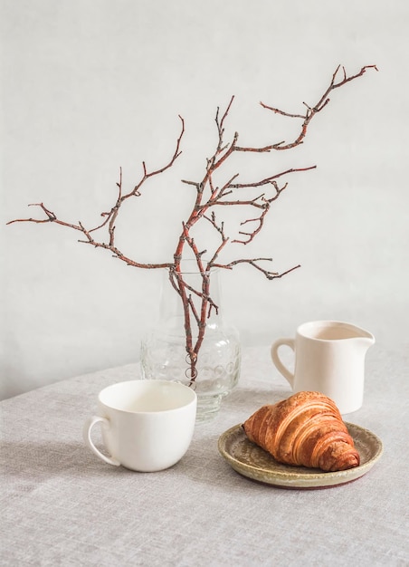 스칸디나비아 홈 인테리어 크루아상 꽃병에는 나뭇가지 우유 주전자와 컵이 밝은 아늑한 방의 탁자에 있습니다.