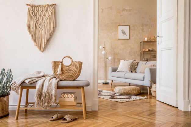 Interni scandinavi di open space con panca in legno, divano grigio, cuscini, palido, cornice per foto, macramè, piante, libri, tappeti, decorazioni ed eleganti accessori personali nell'arredamento della casa.