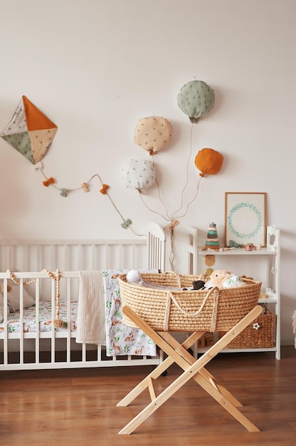 Scandinavian style white interior children's room, bedroom, nursery. Baby cot with canopy. Wooden shelves and toys. Wicker cradle with legs. Textile kite and balloons.