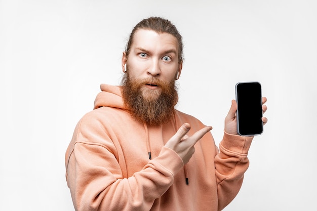 Scandinavische knappe verraste man met een telefoon en wijst naar zwart schermmodel met koptelefoon geïsoleerd op een grijze achtergrond Rustige man met gemberkapsel en baard Moderne digitale technologieën