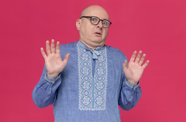 Scared adult slavic man in blue shirt wearing glasses standing with raised hands 