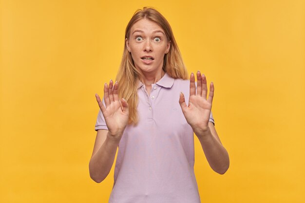 Scared afraid woman with freckles in lavender tshirt keeps hands on front of herself and defending from enemies on yellow