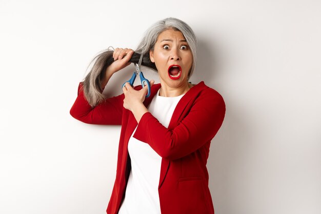 Scared asian senior woman cutting hair with scissors and screaming in panic, standing over white background