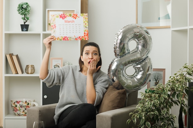 Bocca coperta spaventata con la mano bella donna il giorno delle donne felici che tiene il calendario seduto sulla poltrona in soggiorno