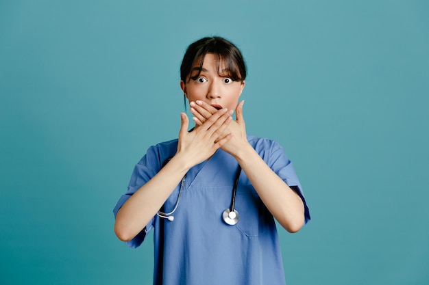 青い背景で隔離の均一なフィス聴診器を身に着けている若い女性医師の手で覆われた口を怖がらせる