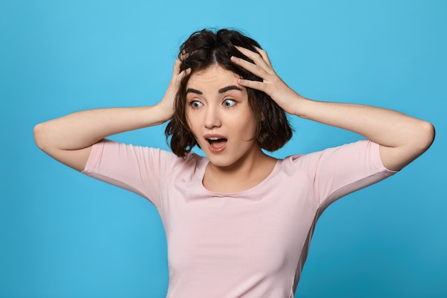 Scared curly young brunette woman on blue background. Human emotions, facial expression concept.