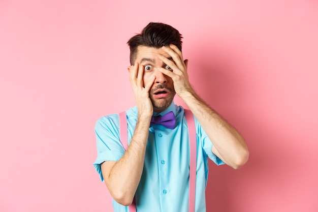 Scared and embarrassed nerdy guy in bow-tie covering his eyes, peeking through fingers at something scary, standing on pink background.