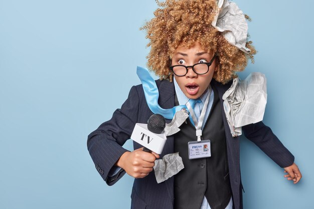 Scared frightened female news presenter holds microphone wears\
spectacles formal clothing reacts to something horrible prepares\
for live broadcasting isolated over blue background journalism\
concept