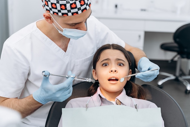 Ragazza spaventata su una poltrona del dentista, impaurita dall'iniezione. il dentista tiene in mano una siringa con un ago.