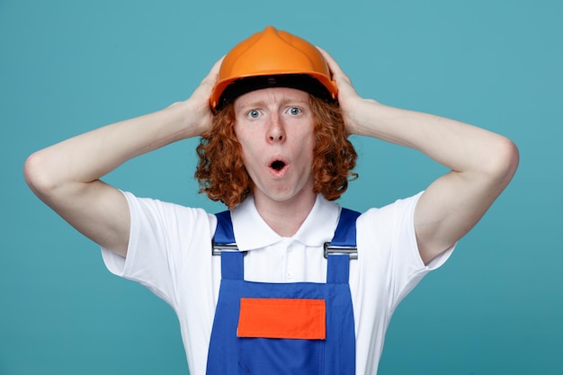 Scared grabbed head young builder man in uniform isolated on blue background