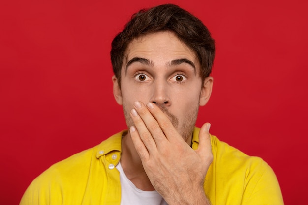 scared handsome man in yellow shirt covering mouth with hand, stares at camera, has bated breath, isolated on red  background