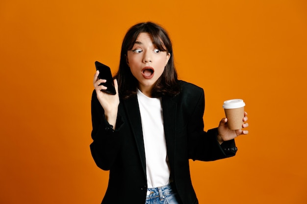 Scared holding coffee cup with phone young beautiful female wearing black jacket isolated on orange background