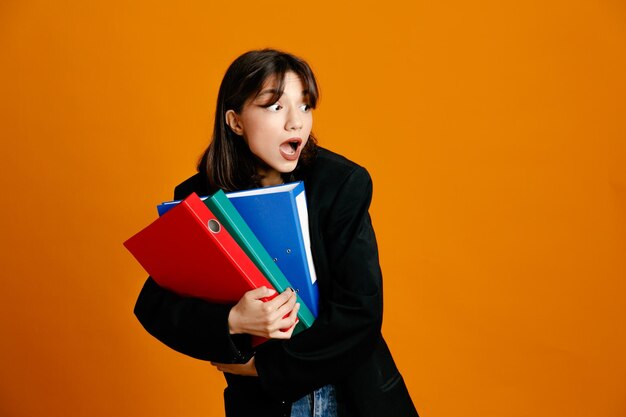 Scared holding folders young beautiful female wearing black jacket isolated on orange background