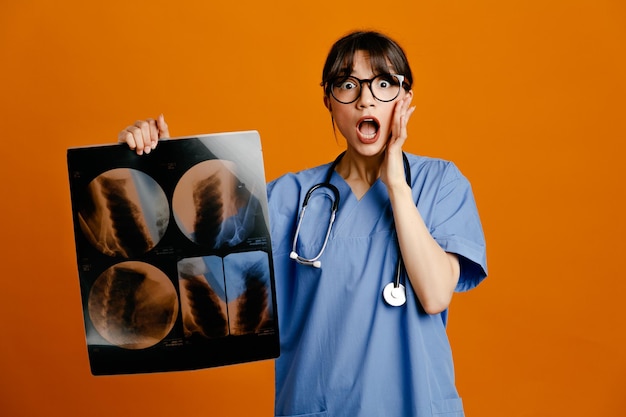 Scared holding xray young female doctor wearing uniform fith stethoscope isolated on orange background