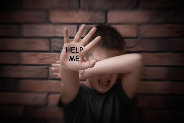 Photo scared little boy with text help me on his hand near brick wall