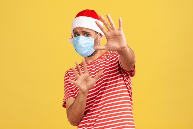 Scared man in medical protective face mask in red santa claus hat holding hands raised afraid of covid2019 virus during holidays shock and panic Indoor studio shot isolated on yellow background