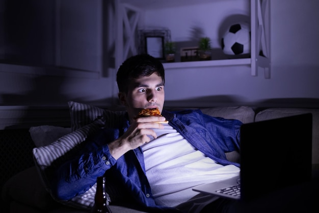 Scared man with laptop and pizza, watching horror movie at night