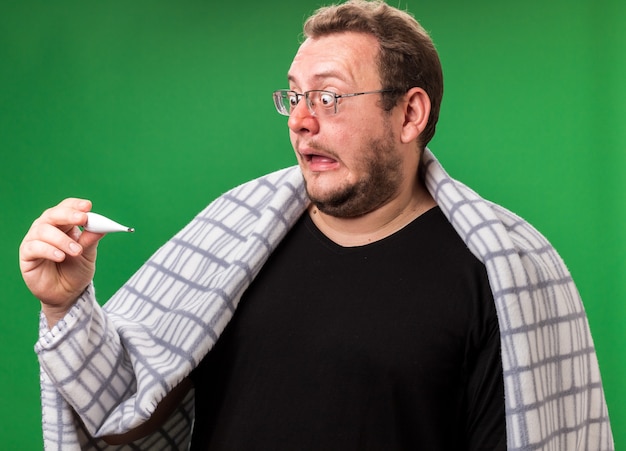 Photo scared middle-aged ill male wrapped in plaid holding and looking at thermometer