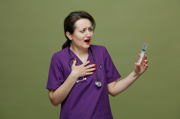 Scared middleaged female doctor wearing uniform and stethoscope around neck holding syringe with needle looking at it while keeping hand on chest isolated on olive green background