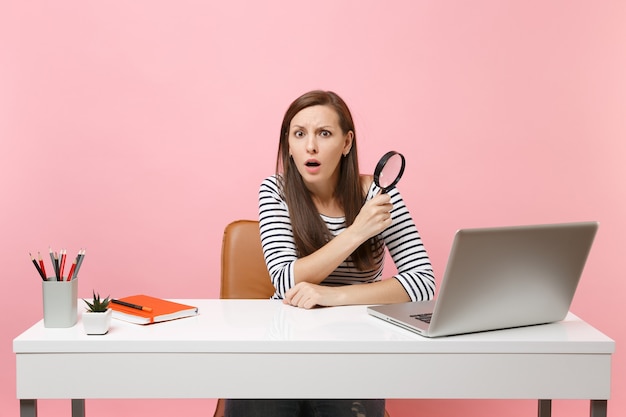 Scared puzzled woman in perplexity holding magnifying glass sitting working on project at white desk with pc laptop isolated on pastel pink background. Achievement business career concept. Copy space.