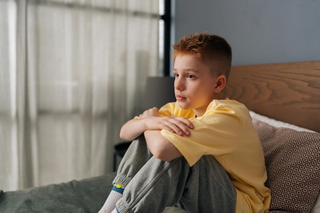 scared sad little boy sitting on bed alone with hugging knee and sadness looking away feeling pressure and problem thoughtful stressed child in bedroom
