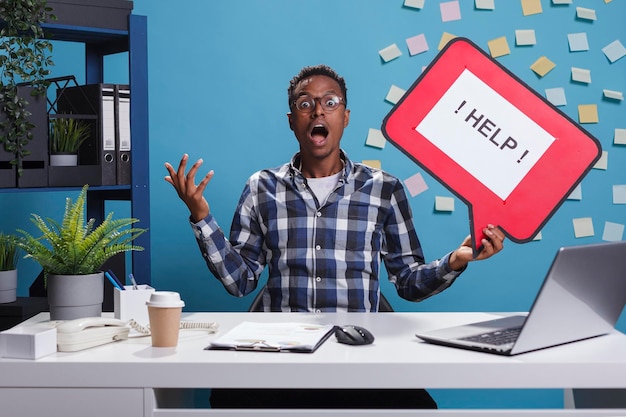 Scared screaming business man hodling cardboard bubble sign requesting help from coworkers because of danger. Shocked and afraid young adult entrepreneur calling for help while at desk in office.