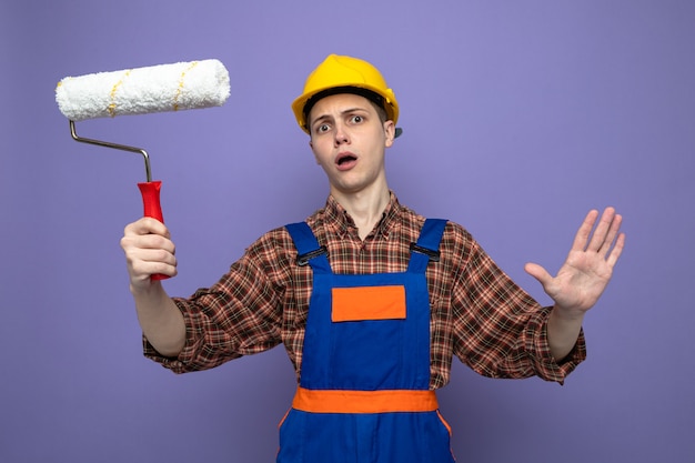 Scared showing stop gesture young male builder wearing uniform holding roller brush 