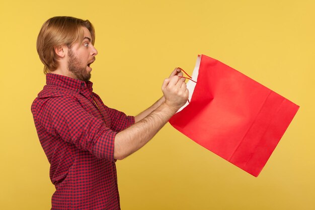 Scared surprised shopper guy in checkered shirt looking inside shopping bag with big eyes open mouth in amazement, shocked by purchase. indoor studio shot isolated on yellow background, side view