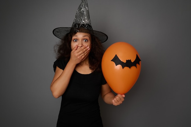 Scared woman dressed in Halloween witch costume, covering her mouth in fear and horror, looking at camera, posing with colorful balloons with felt-cut black bat against gray background with copy space