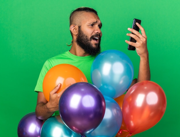 Scared young afro-american guy wearing green t-shirt standing behind balloons holding and looking at phone 