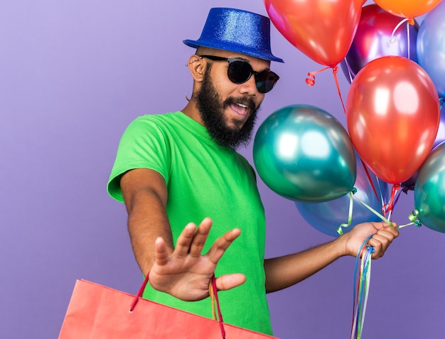 Испуганный молодой афро-американский парень в партийной шляпе в очках держит воздушные шары с подарочной сумкой, показывая жест остановки, изолированный на синей стене