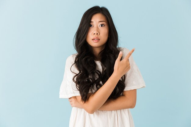 Испуганная молодая азиатская красивая женщина позирует изолированной над синей стеной, указывая
