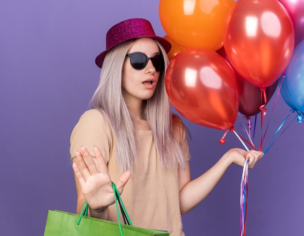 Scared young beautiful girl wearing party hat with glasses holding balloons with gift bag showing stop gesture 