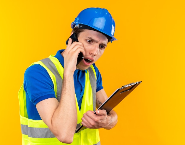 Scared young builder man in uniform holding clipboard speaks on phone 